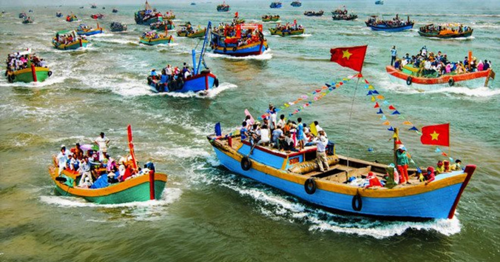 Đoàn rước kiệu Cá Ông đi qua làng chài ven biển, người dân mặc áo dài truyền thống, cờ phướn rực rỡ trong nắng sớm.