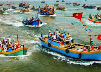 Đoàn rước kiệu Cá Ông đi qua làng chài ven biển, người dân mặc áo dài truyền thống, cờ phướn rực rỡ trong nắng sớm.