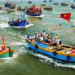 Đoàn rước kiệu Cá Ông đi qua làng chài ven biển, người dân mặc áo dài truyền thống, cờ phướn rực rỡ trong nắng sớm.
